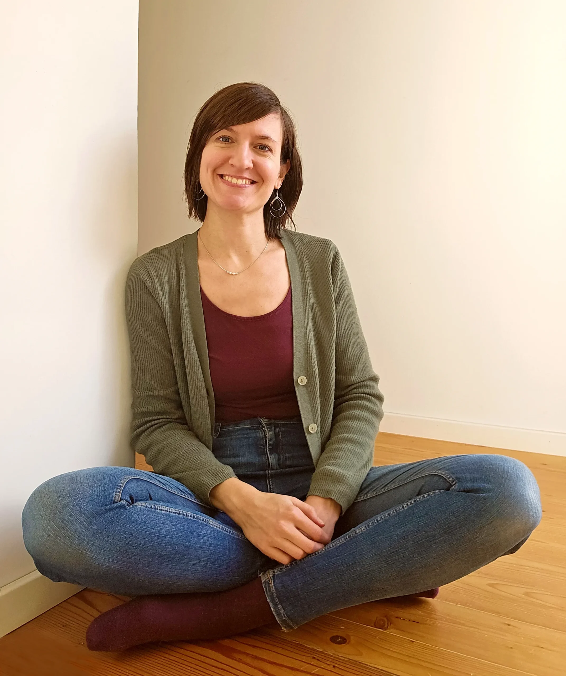 Porträt einer Klinischen Psychologin, die lächelnd im Schneidersitz auf einem Holzboden vor einer weißen Wand sitzt – persönliche Vorstellung auf der Startseite einer psychologischen Praxis in Villach und online