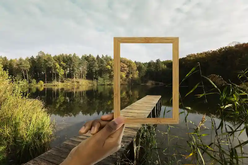 Supervision – Reflexion in einem professionellen Rahmen | Villach & Online Hand hält einen leeren Holzrahmen vor eine ruhige Seenlandschaft mit Steg – Supervision als professioneller Rahmen zur Reflexion beruflicher Themen bei Klinischer Psychologin in Villach und online